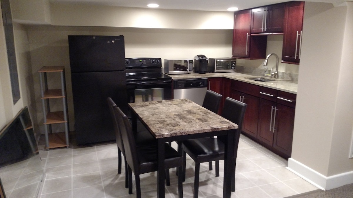 A compact kitchen area is shown, featuring dark wood cabinets and a modern black appliance set, including a refrigerator, stove, and microwave. A small dining table with four chairs occupies the space, complemented by a light-colored countertop.
