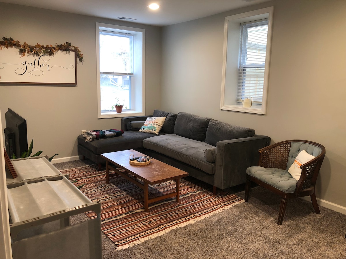 A living room area features a large sectional sofa with a chaise, accompanied by a coffee table on a patterned rug. Two windows allow natural light to brighten the space. A television sits across from the seating, with a decorative wall sign above.