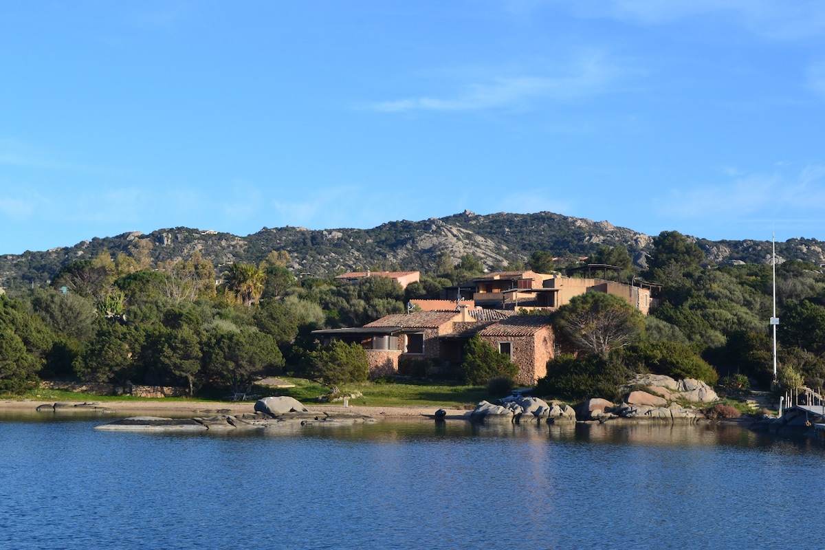 A serene view of a villa situated among lush greenery and rocky formations along a tranquil body of water. The structure features warm-toned exterior walls and is set against a backdrop of gently rolling hills under a clear blue sky.
