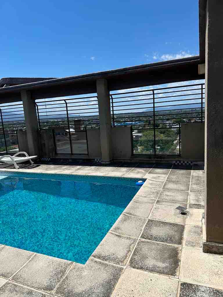 A rooftop pool area is visible, featuring clear water surrounded by light-colored tiles. Lounge chairs are positioned nearby, and a panoramic view of the city can be seen through the protective glass railing.