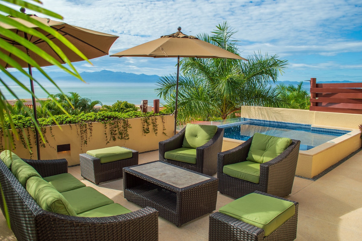 An outdoor seating area is nestled among lush greenery, featuring a comfortable arrangement of dark wicker furniture with green cushions. A small table is positioned at the center, while large umbrellas provide shade. In the background, a private pool reflects the scenic view of the ocean.