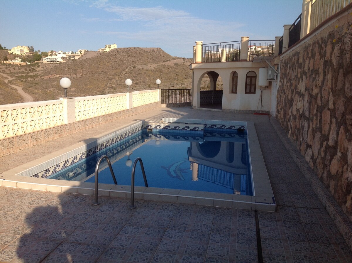 A private swimming pool is situated on a sunlit terrace, surrounded by a tiled area and stone walls. The water reflects the clear sky above. Two steps lead into the pool, with lounge space available on the surrounding deck.