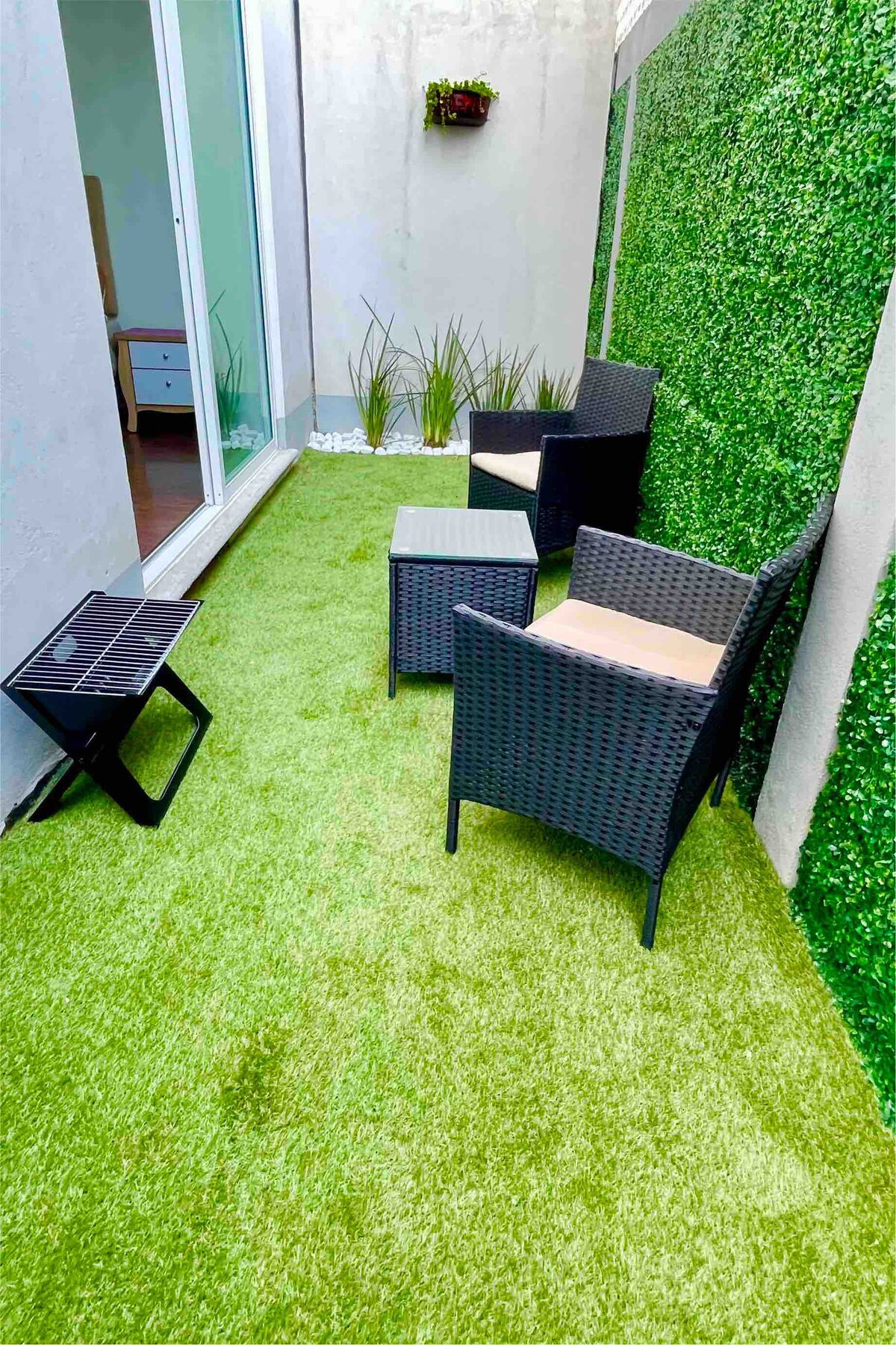 A private garden area features artificial grass, accompanied by two wicker chairs and a small table. A compact grill is positioned nearby. Lush green wall coverings enhance the serene outdoor space, while large glass doors provide access to the interior.