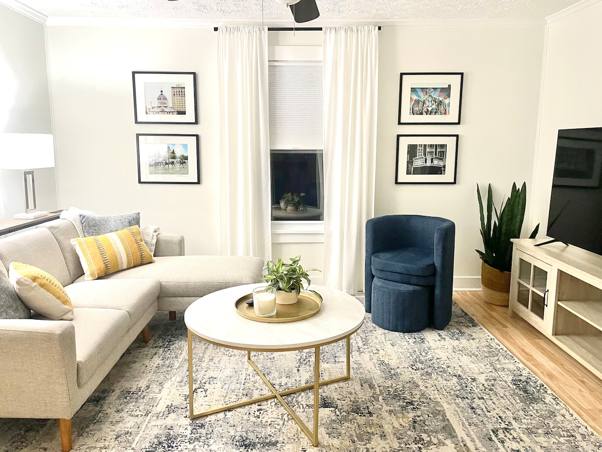 A comfortable living area features a sectional sofa adorned with colorful cushions and a round coffee table at its center. Framed photographs are displayed on the walls, while light curtains frame the window. A blue accent chair and a potted plant complete the inviting space.