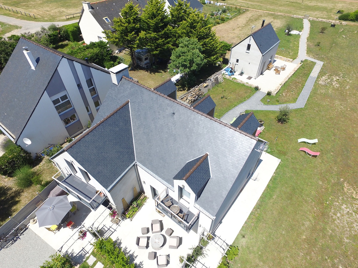An aerial view captures a contemporary house surrounded by greenery and outdoor spaces. Terraces are visible with seating arrangements and pathways leading to the garden. The proximity of the house to open fields can be observed, providing a tranquil setting.