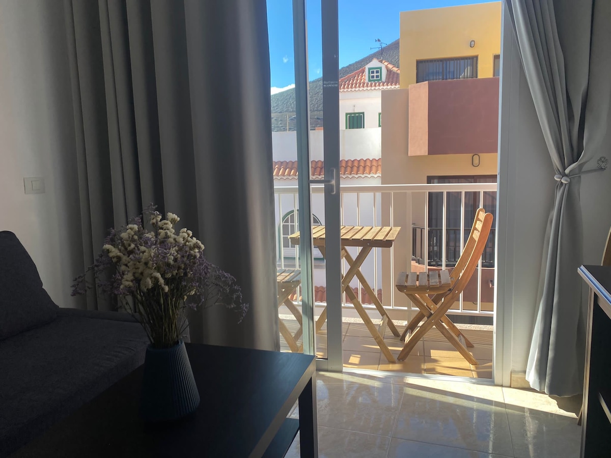 A light-filled space features glass doors opening to a small balcony. Two wooden chairs and a table are set on the balcony, offering views of buildings and mountains in the background. Soft curtains frame the view, enhancing natural light within the room.
