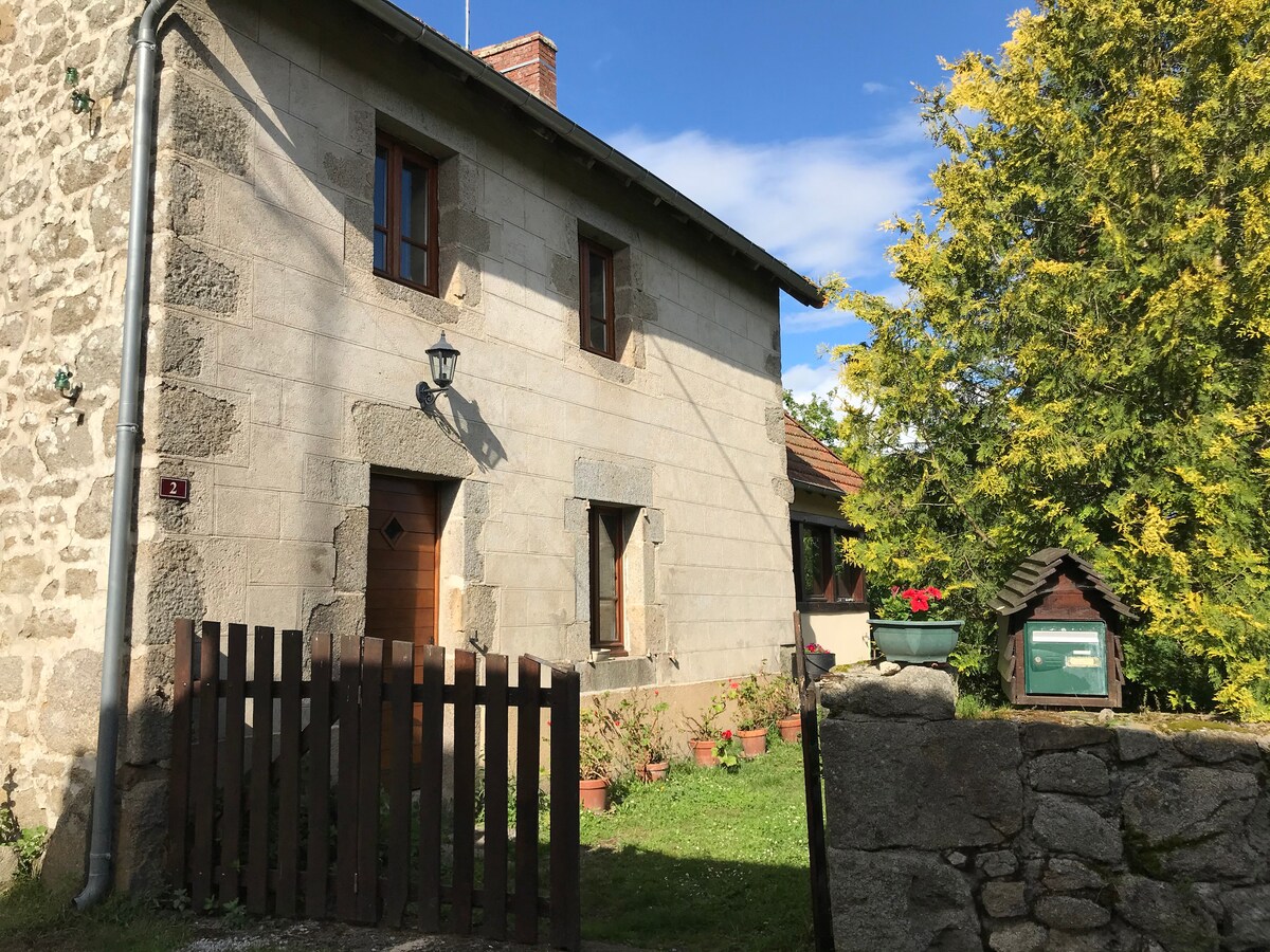 A charming two-story stone house is presented, featuring a warm grey facade with traditional windows. A wooden fence lines the front, complemented by a well-maintained green lawn and flower pots. An inviting pathway leads to the entrance, surrounded by nature.