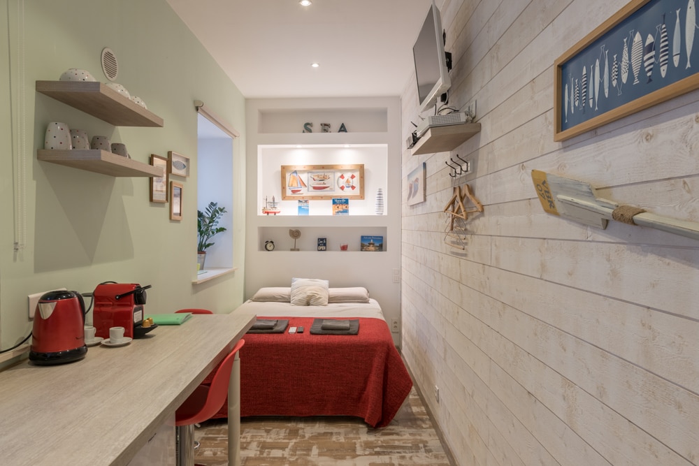 A mini studio features light green walls and wooden paneling. A cozy sleeping area is seen with a bed draped in red bedding. Shelves hold decorative items, while a kitchenette is equipped with a coffee machine. A small dining table and wall-mounted TV enhance the space.