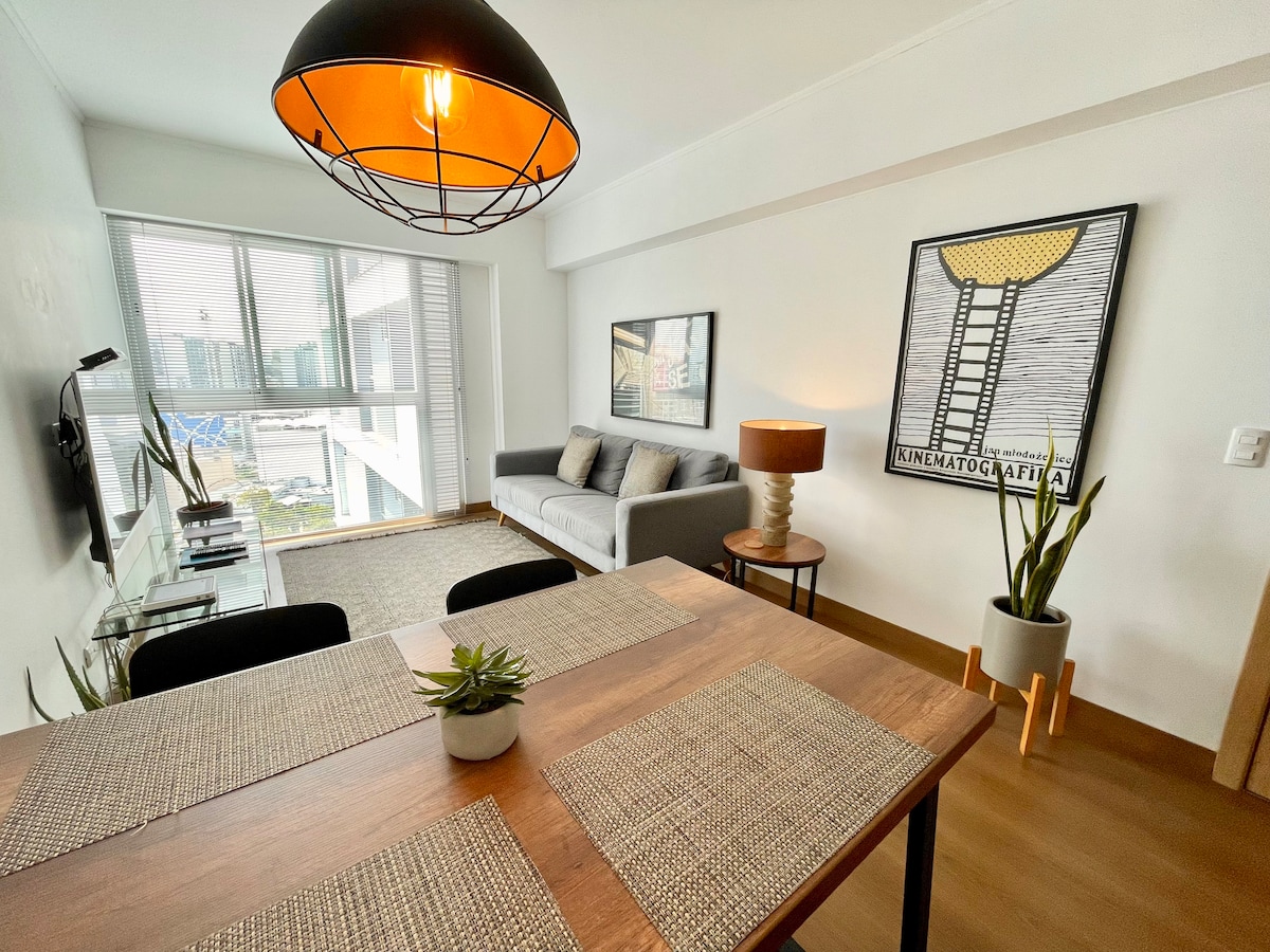 A modern dining area features a wooden table with place settings and a decorative plant. A cozy living space includes a light grey sofa and a side lamp, complemented by large windows allowing natural light and views of the surroundings.