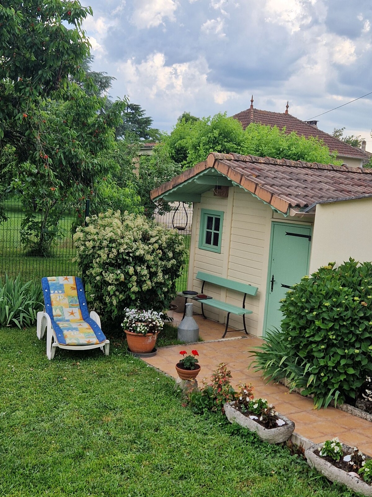 A charming exterior view showcases a small building surrounded by lush greenery. A vibrant lounge chair is positioned on a patio alongside flower pots and a decorative stone border, enhancing the garden's natural beauty. The building features light-colored walls and a green door, inviting relaxation.