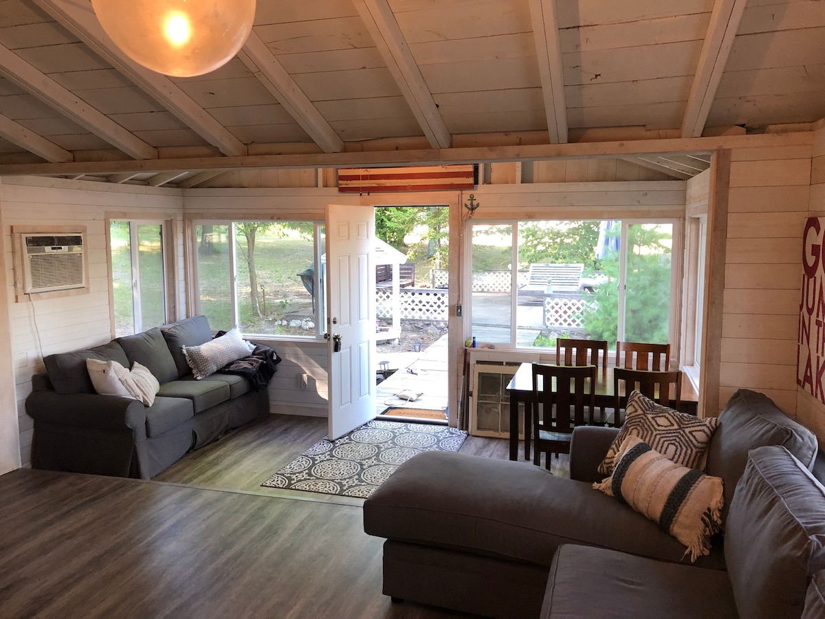 The main living area features two comfortable sofas and a dining table, providing a gathering space that is filled with natural light. Large windows overlook the outdoor area, highlighting views of the surrounding nature and facilitating easy access to the entrance.