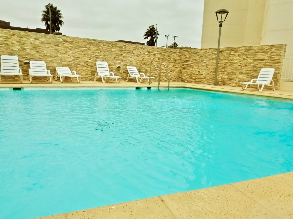 A clear swimming pool is surrounded by light-colored tiles and features several white lounge chairs positioned for relaxation. A low stone wall provides privacy, while palm trees are visible in the background under an overcast sky.