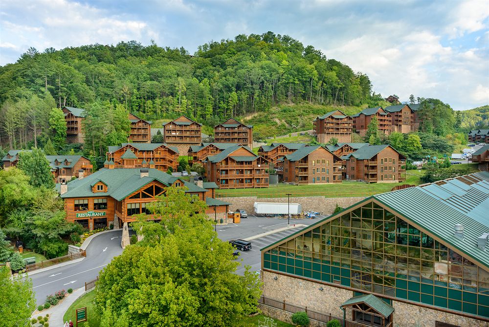 A panoramic view of the Westgate Smoky Mountain Resort showcases a collection of rustic wooden buildings nestled among lush green hillside. The clear blue sky complements the natural setting, while the vibrant foliage enhances the mountain retreat atmosphere.