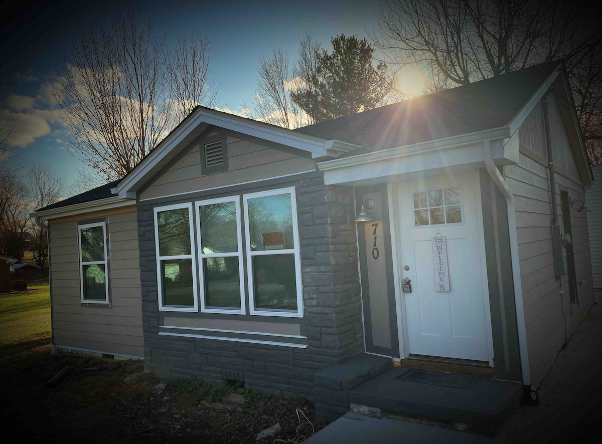 The exterior of the house is visible, showcasing a combination of stone and siding materials. Large windows allow natural light to brighten the front entry, where a white door leads inside. A landscaped front yard features small trees and an open grassy area.