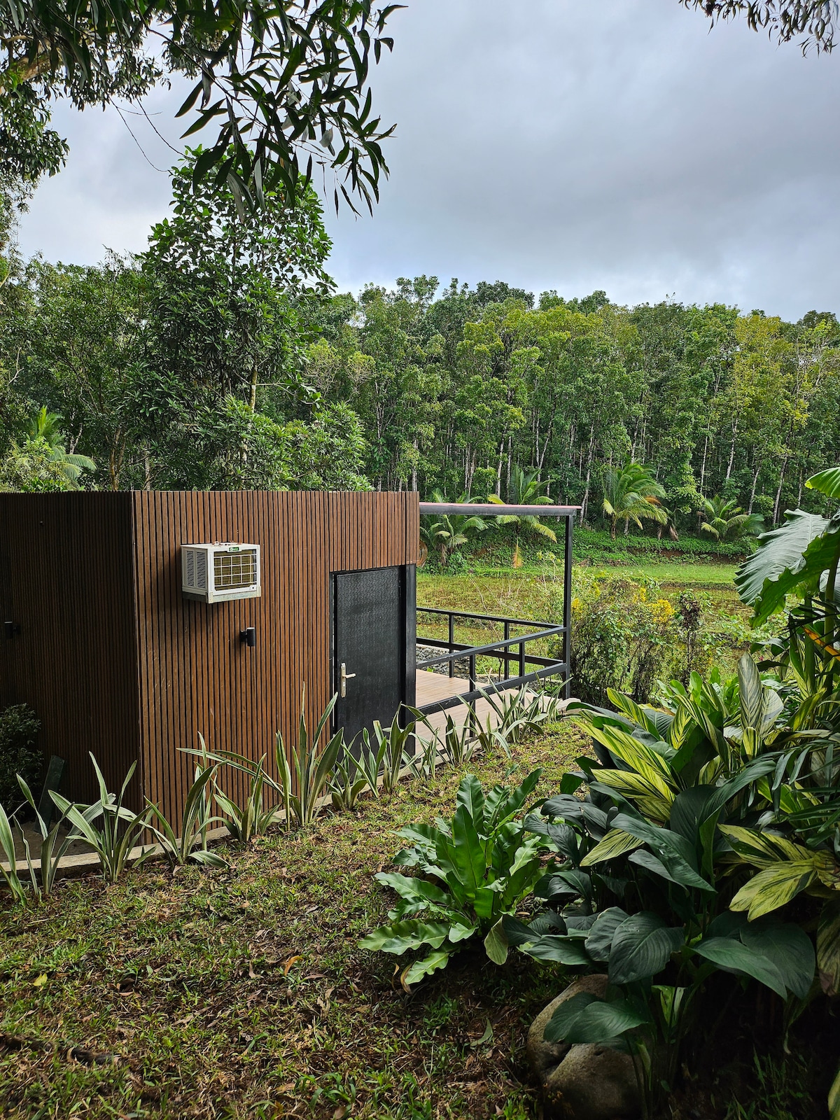 A riverside cabin is set amidst lush greenery, featuring a wooden exterior and an air conditioning unit on the side. A pathway leads to a private patio, which offers a view of surrounding trees and nearby rice fields, contributing to a serene atmosphere.