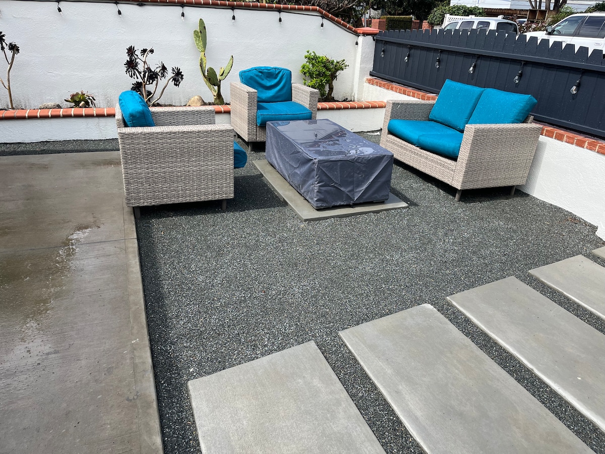 An outdoor seating area is featured, consisting of two light-colored wicker armchairs and a matching loveseat, all adorned with bright blue cushions. A rectangular table with a protective cover is positioned in the center, surrounded by pebbled ground and bordered by low-growing plants.