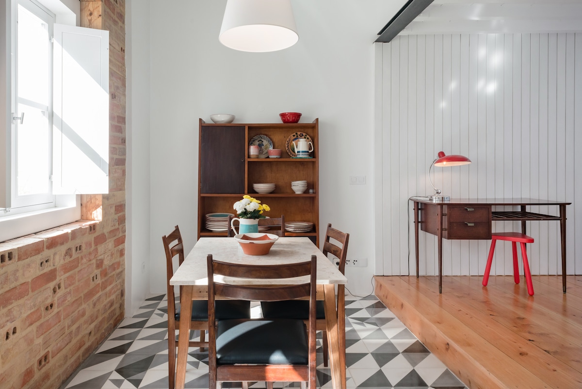 A modern dining area is highlighted by a wooden table surrounded by six chairs. Natural light floods in through a window, illuminating the textured brick wall. A display shelf filled with plates complements a small workspace featuring a vintage desk and a bright red chair.