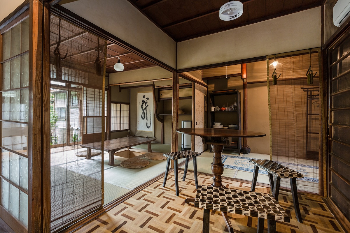 A serene dining area features a wooden table surrounded by intricately designed chairs. Sliding shoji screens separate the space from tatami mat rooms, creating an inviting transition to the connected garden. Natural light fills the room, highlighting the traditional wooden flooring and decor.