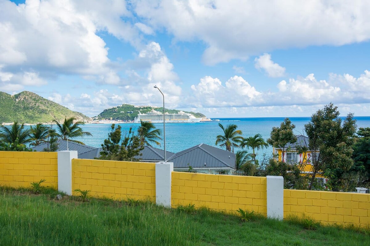 An ocean view is presented, showcasing vibrant blue waters under a partly cloudy sky. A yellow wall borders the foreground, with lush green grass and palm trees adding natural elements. In the background, rolling hills and residential structures are visible, enhancing the coastal atmosphere.