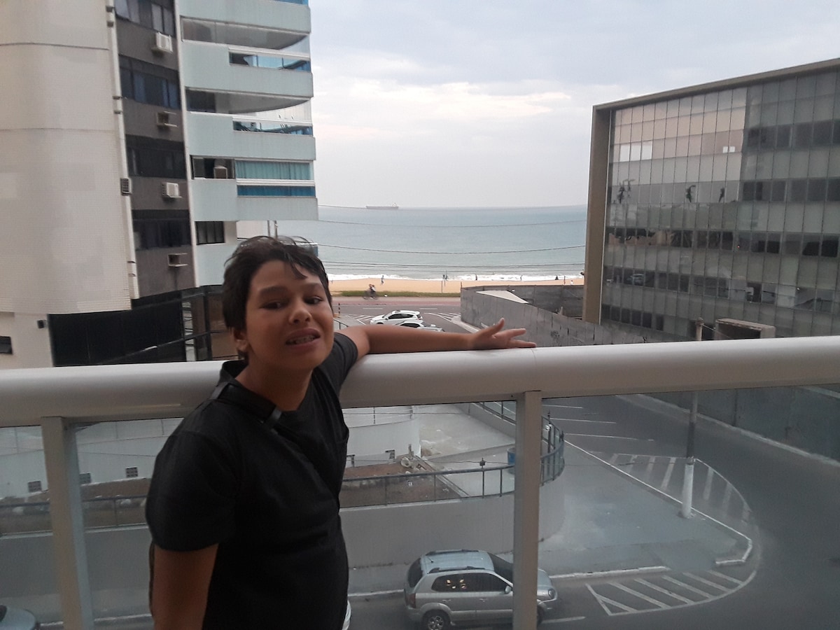 A view from an enclosed balcony showcases the ocean horizon and sandy beach in the distance. The surrounding buildings are visible, with a street and parked vehicles below, creating a sense of urban ambiance.