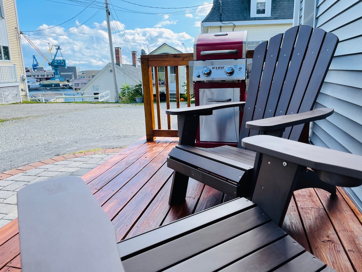 The farmers porch features two comfortable adirondack chairs placed on warm wooden decking. A BBQ grill is positioned nearby, with a backdrop of distant harbor views under a blue sky. The scene is designed for outdoor relaxation and enjoyment.