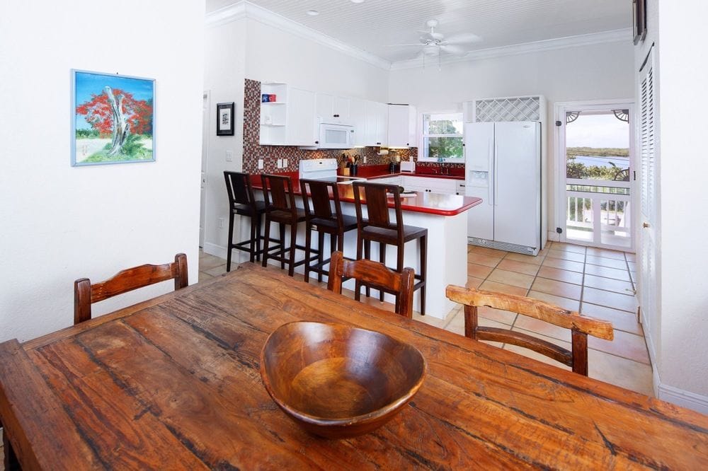 A dining area features a rustic wooden table with several chairs surrounding it, providing a cozy space for meals. In the background, a modern kitchen is visible, equipped with white cabinetry and appliances, including a refrigerator and stove. Light permeates through a window, enhancing the inviting ambiance.