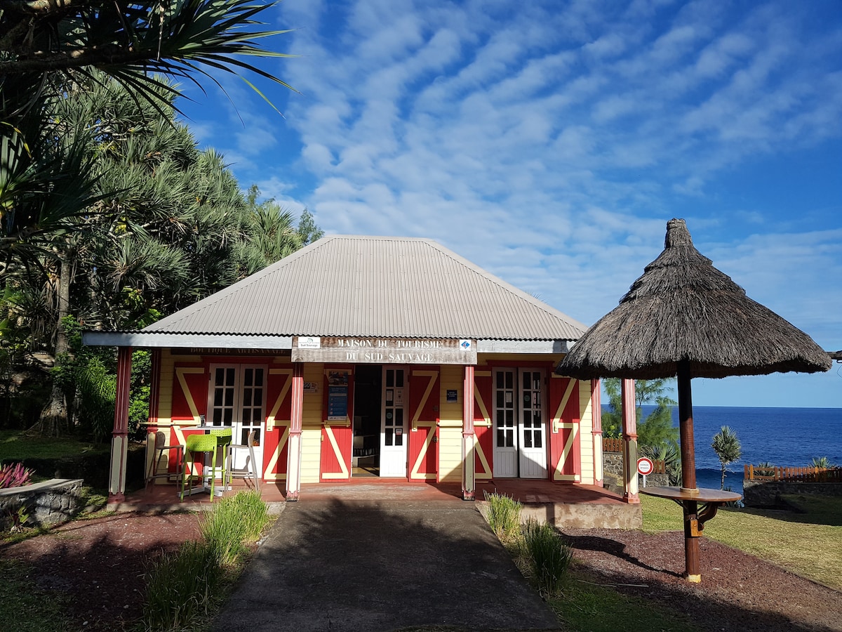 A charming building is positioned near the coast, featuring a peaked roof and a bright red and yellow exterior. Lush greenery surrounds the front, complemented by a thatched pavilion nearby, offering a scenic view of the ocean and sky.