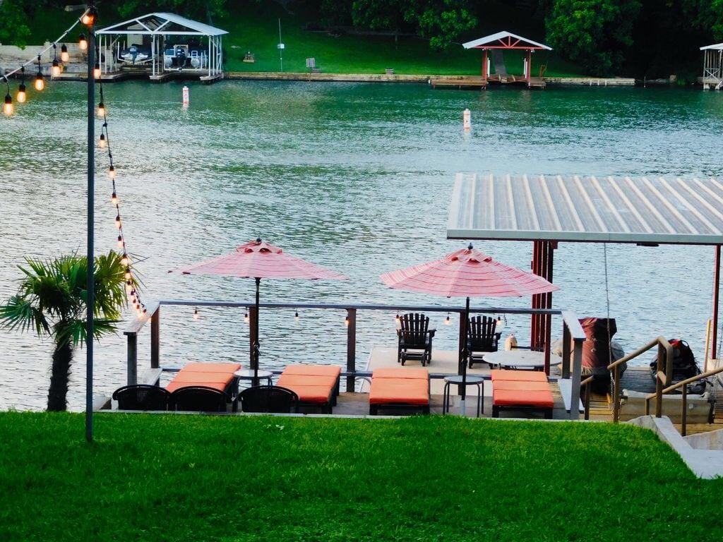 An outdoor lounge area is situated by the water, featuring orange lounge chairs beneath red umbrellas. A dock and two covered boat slips are visible in the background, surrounded by lush greenery and string lights that add a relaxed ambiance.