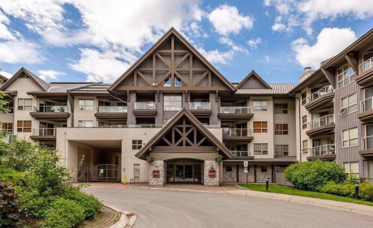 The exterior of a well-maintained condo building is showcased, featuring a combination of modern architecture and natural landscaping. Large windows allow ample natural light, and a grand entrance with decorative elements is accentuated by greenery on either side.