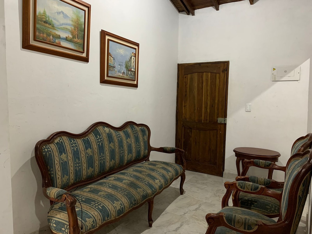 A cozy seating area features a patterned sofa accompanied by two matching armchairs. A pair of small wooden tables is positioned nearby. Art pieces depicting landscapes adorn the walls, and a wooden door is visible, leading to another space.