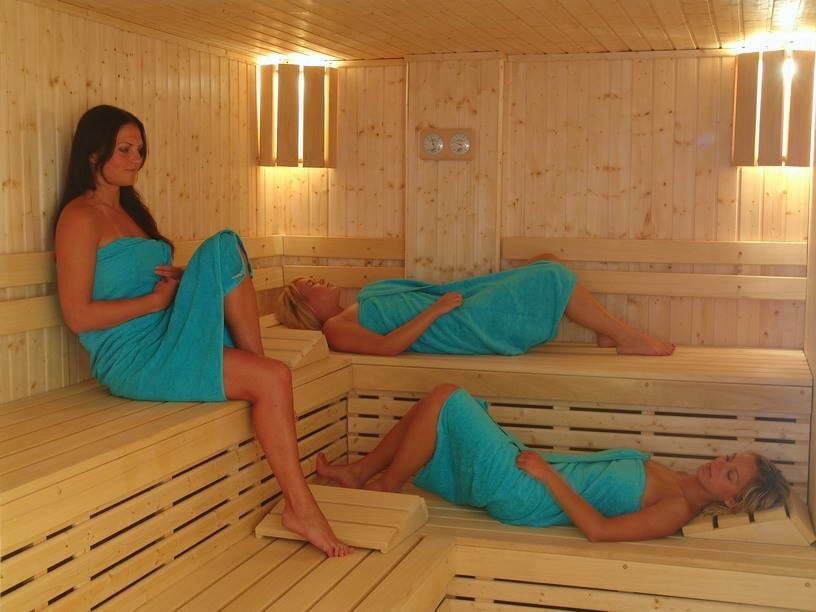 A warm sauna is designed with wooden paneling and seating areas, illuminated by soft lighting. Three individuals are seen relaxing on the benches, wrapped in turquoise towels, creating a serene environment intended for unwinding and rejuvenation.