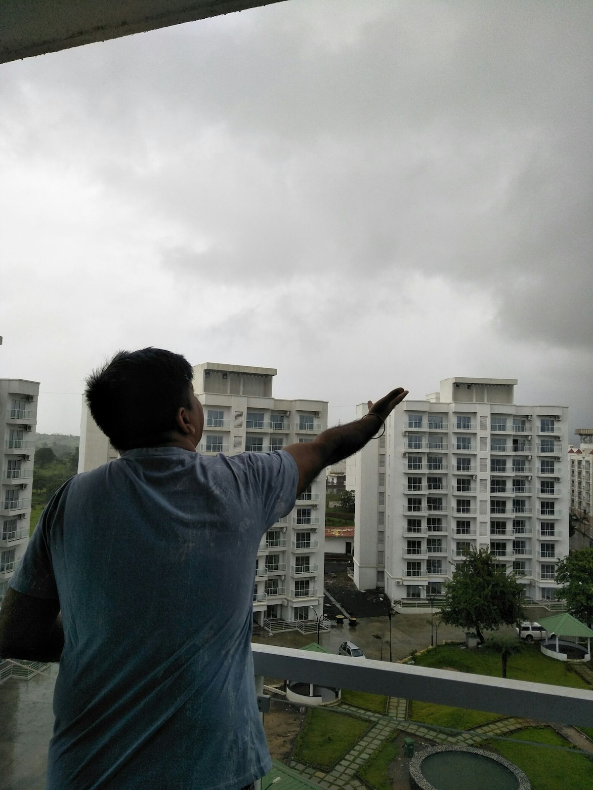 A person stands on a balcony, gazing toward an overcast sky filled with gray clouds. Modern apartment buildings can be seen in the background, surrounded by a landscaped area featuring manicured greenery. The atmosphere appears calm, reflecting a serene moment during rainy weather.
