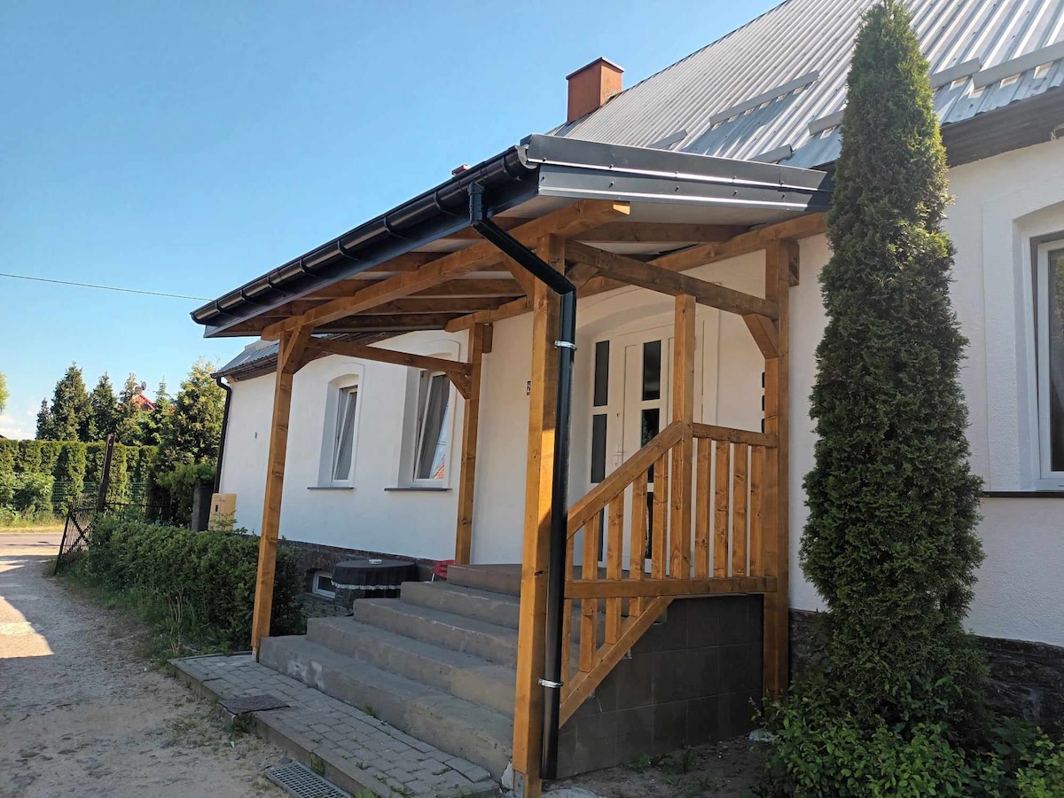 The entrance of the house is highlighted by a wooden porch, featuring a sloped roof and steps leading up to the front door. Side windows provide natural light, and greenery flanks the entrance, contributing to a welcoming exterior.