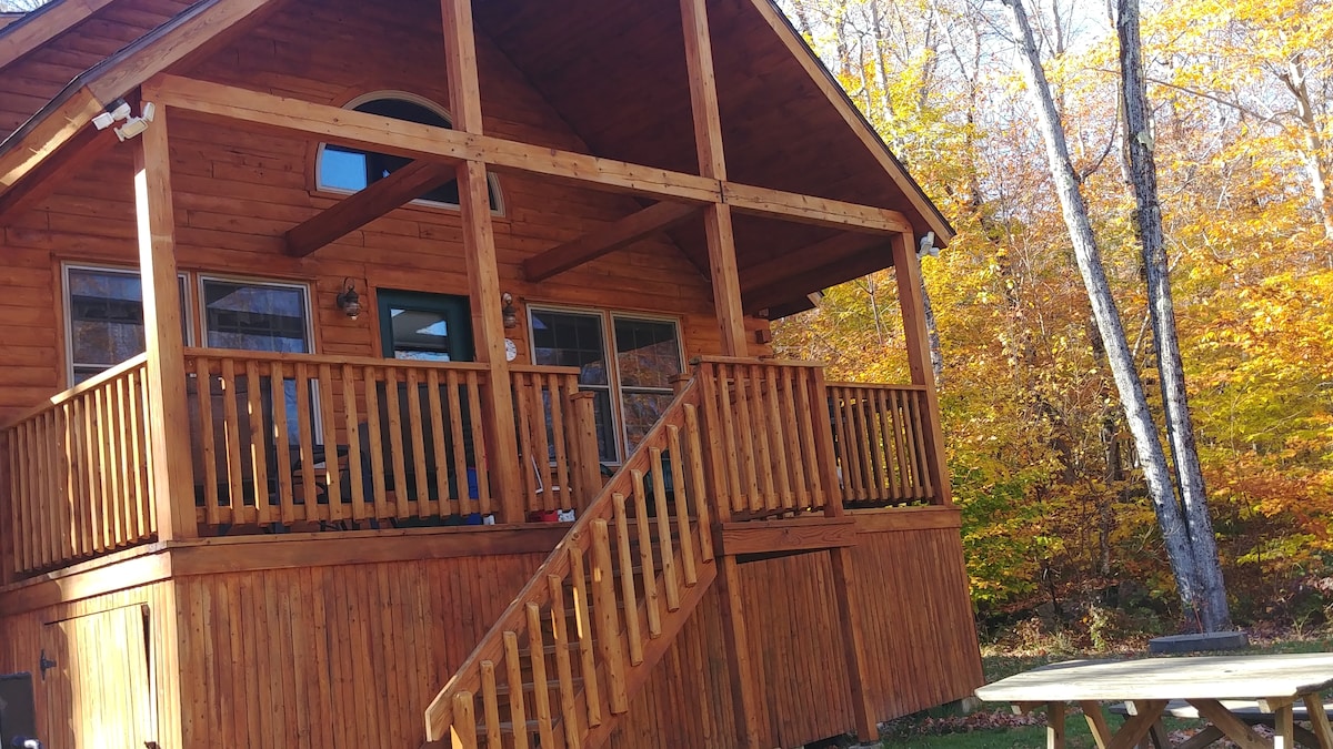 The cabin exterior features warm wooden siding and a covered front porch with a sturdy railing. Two sets of stairs lead up to the main entrance, surrounded by autumn foliage that adds natural color to the setting. A picnic table is seen to the side.