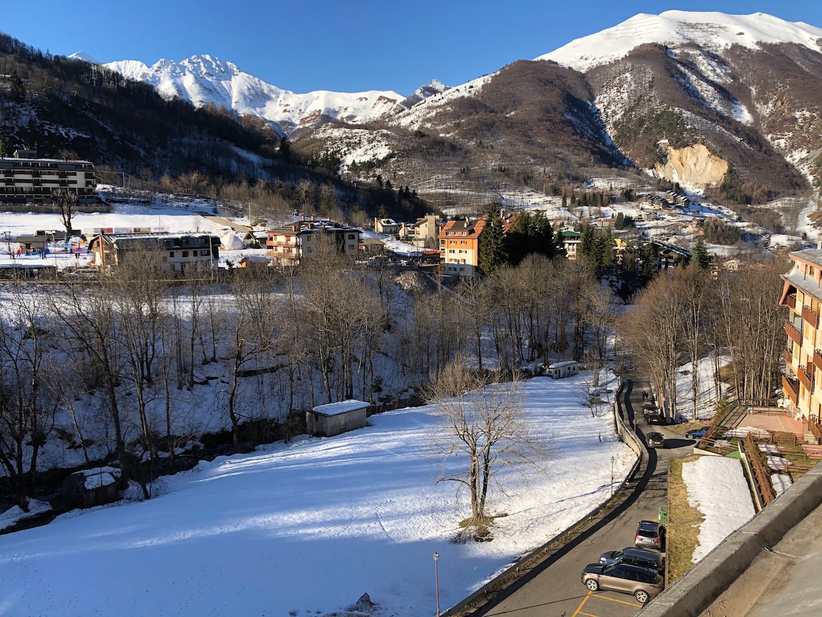 A panoramic view showcases snow-covered mountains and a serene valley. A small road winds through the landscape, lined with bare trees. The scene highlights a combination of residential buildings, with a few vehicles parked beside the road, creating a peaceful winter setting.