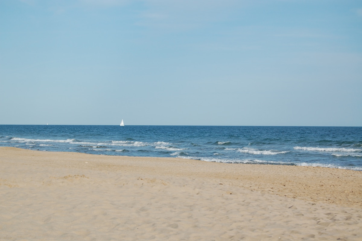 A sandy beach stretches out towards calm waves, with the sea meeting the horizon. A small sailboat is visible in the distance, contributing to a serene coastal view. The clear blue sky provides a bright backdrop, enhancing the tranquil atmosphere.