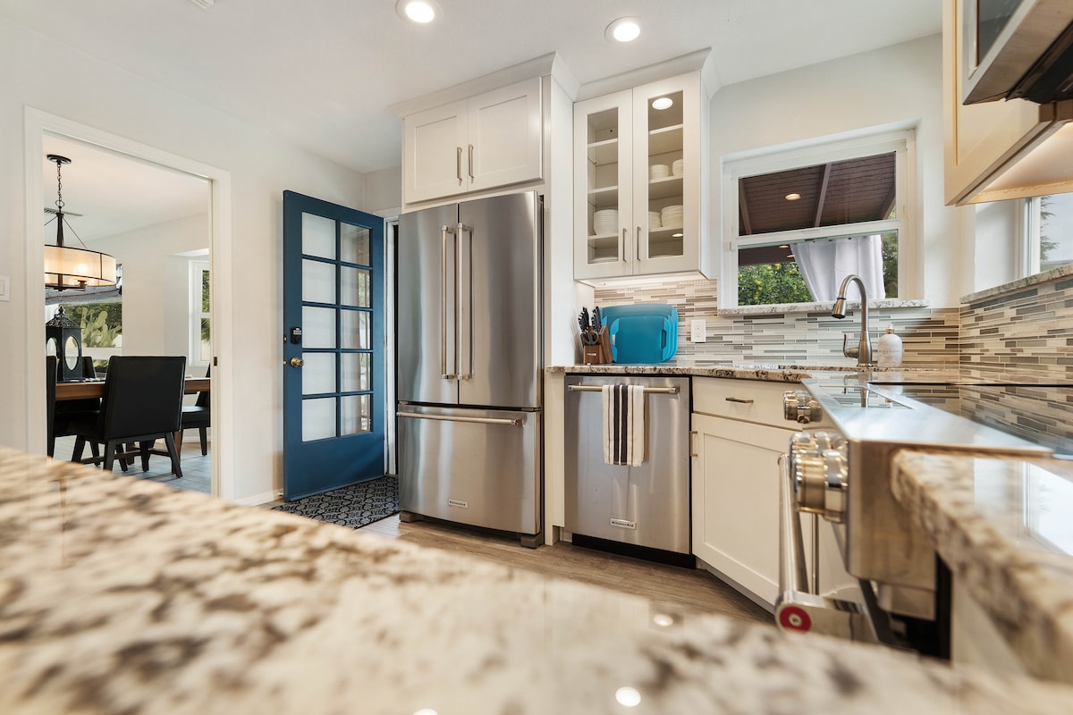 A modern kitchen is showcased, featuring stainless steel appliances, including a large refrigerator and a double sink. Light-colored cabinets contrast with the granite countertops. The window allows natural light, while a blue door adds a pop of color, leading to a welcoming entryway.