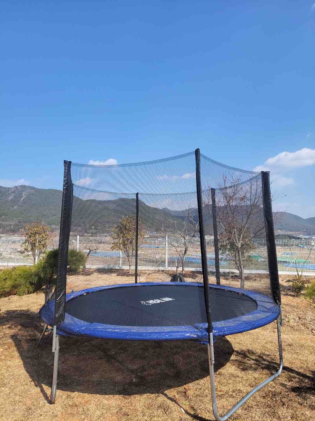 A trampoline is set up in a grassy area, enclosed with a safety net. The surrounding landscape features distant mountains under a clear blue sky, creating an open outdoor space for various activities.