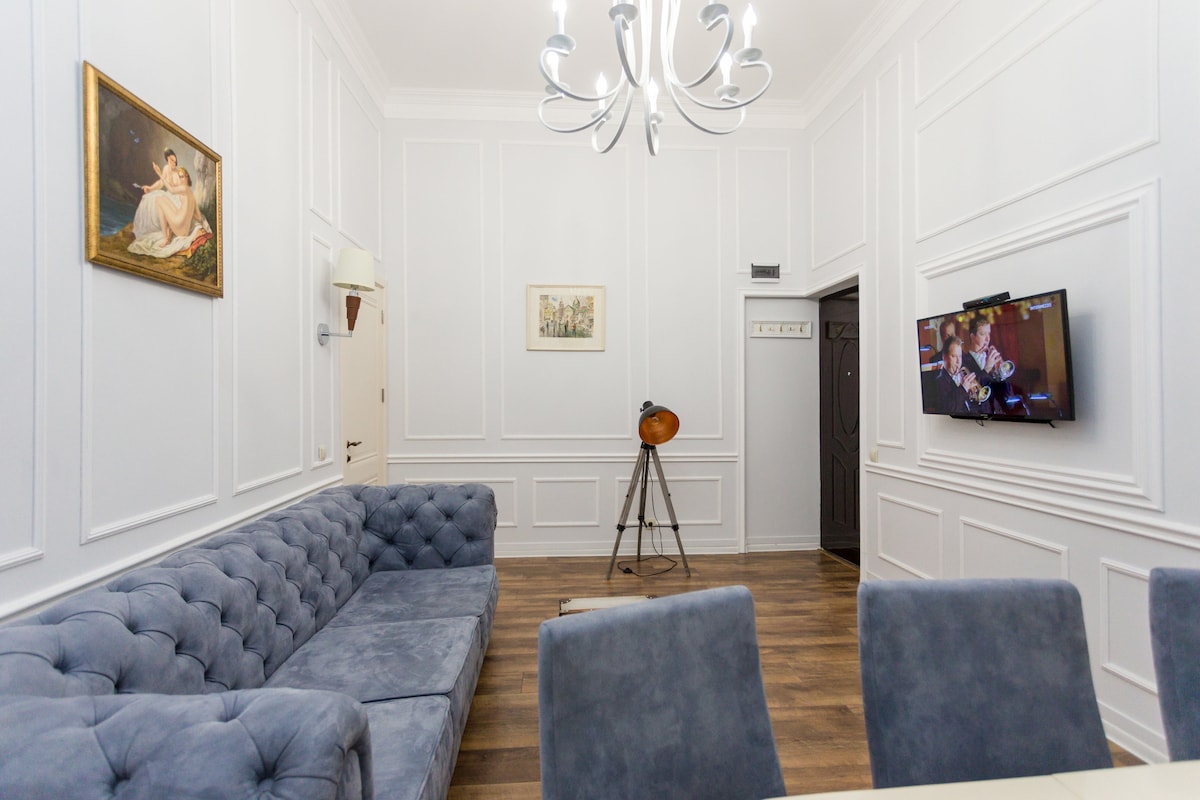 A vintage-style living room features a plush blue sofa and a dining table surrounded by upholstered chairs. A classic chandelier is positioned above, while a TV is mounted on the wall opposite, displaying clear content. Decorative wall paneling enhances the elegant ambiance.