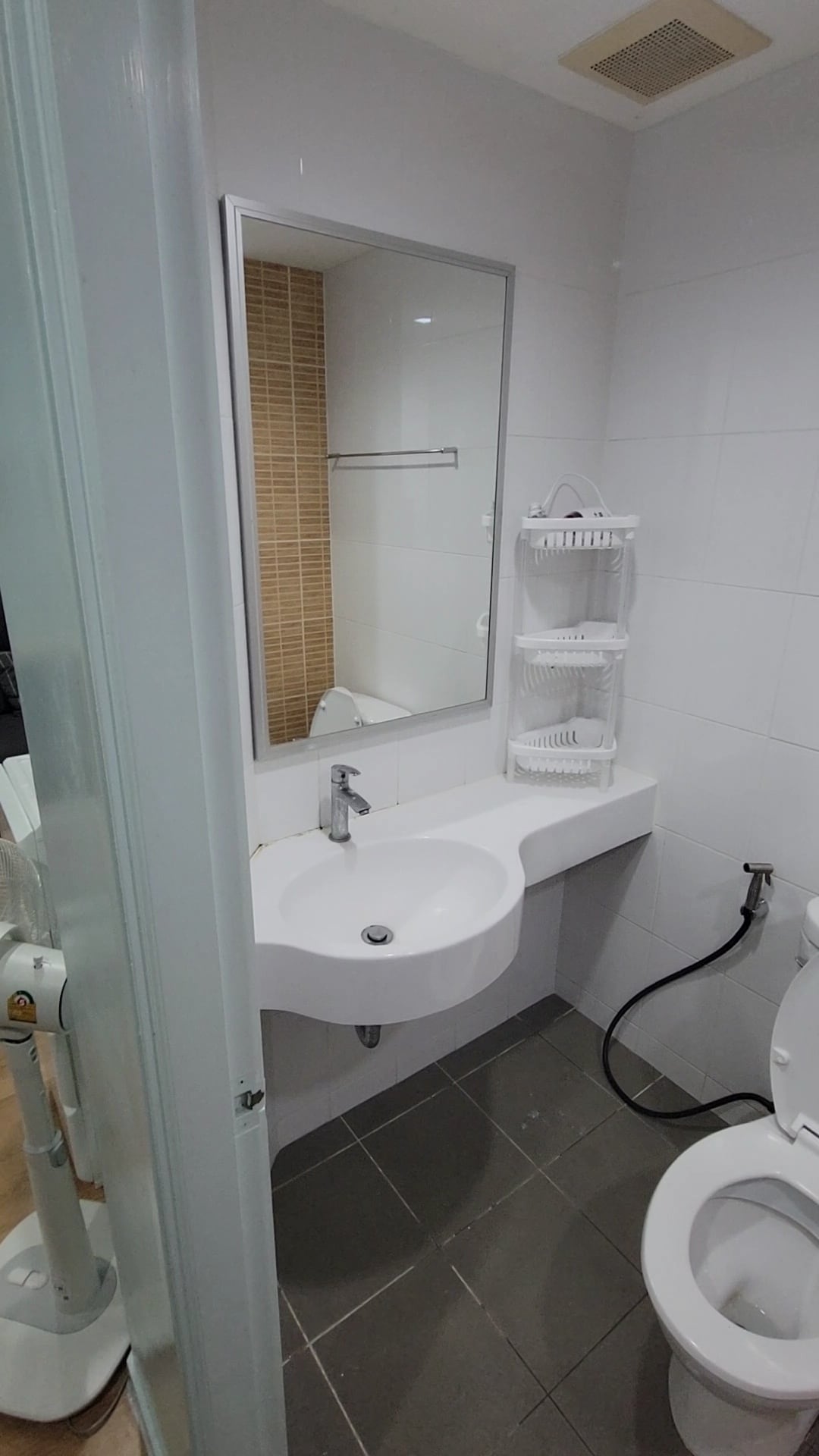 A compact bathroom is presented, featuring a wall-mounted mirror above a curved sink. A toilet is positioned beside the sink, with chrome fixtures visible. Neutral tiles cover the floor, while a shower is partially visible through an open door.