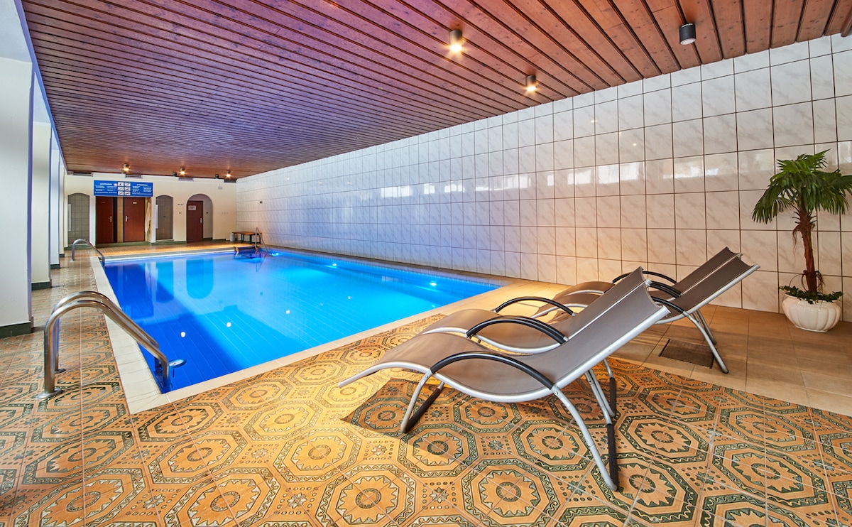 An indoor pool area features a blue swimming pool surrounded by elegant tiles and modern lounge chairs. The ambiance is enhanced by recessed lighting and wood-paneled ceilings, creating a contemporary and relaxing environment.