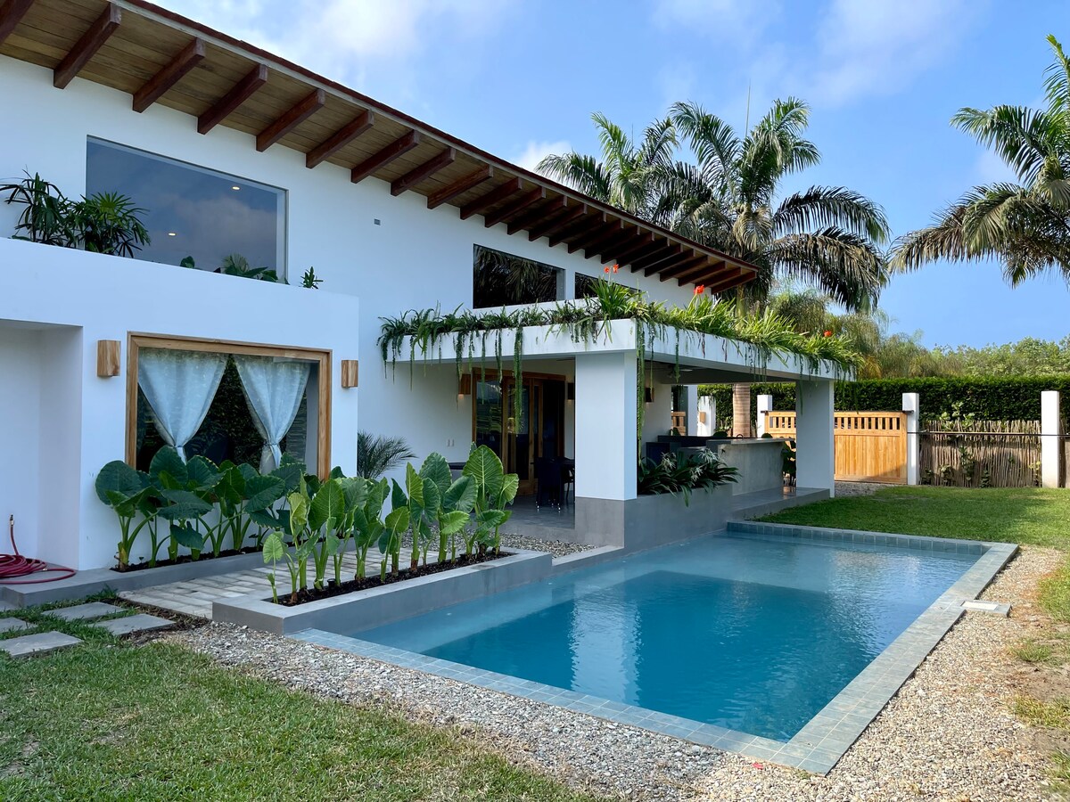 A modern house is showcased, featuring large windows and an inviting terrace with greenery. A pool is positioned in front, accompanied by a spacious lawn and decorative plants. The design harmoniously integrates with the surrounding nature.