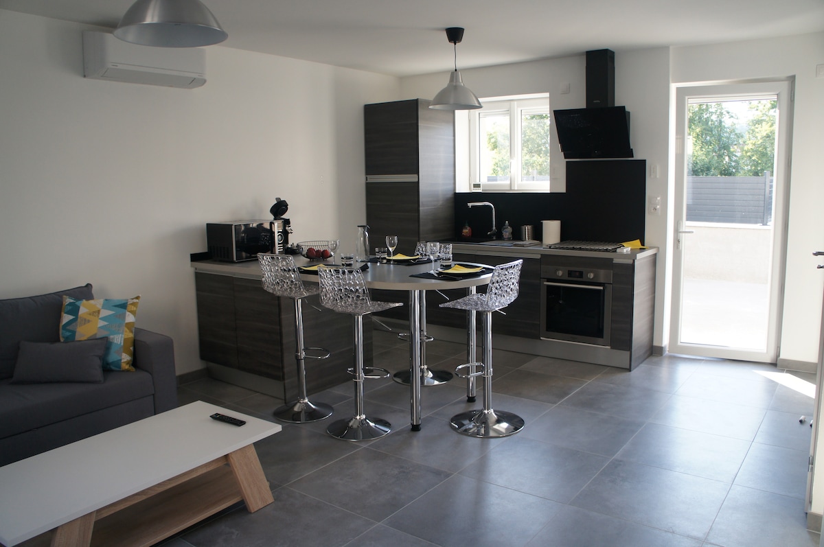 A modern living area features a well-equipped kitchen with sleek cabinetry and a spacious countertop with bar seating. A comfortable gray sofa is positioned near the bright window, with light streaming into the room. A door leads to an outdoor terrace.
