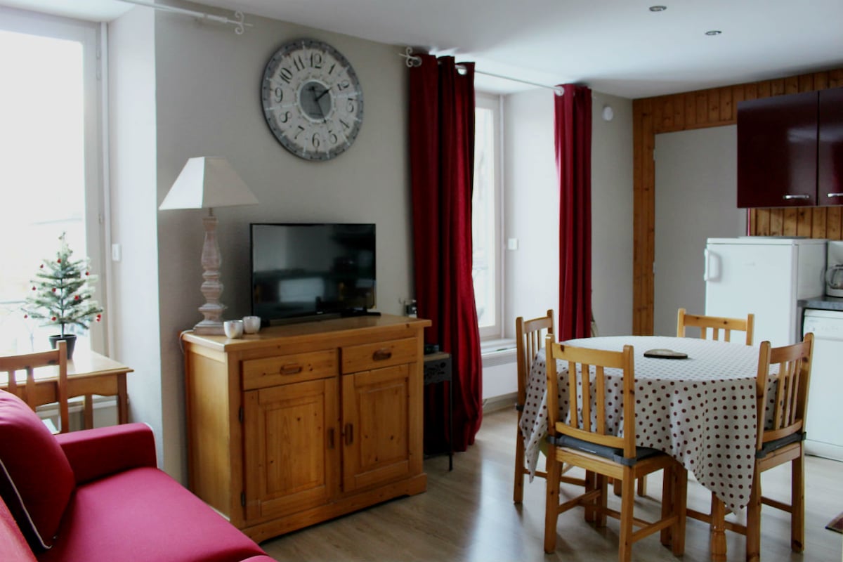 A bright living area features a wooden dining table surrounded by four chairs, complemented by a television on a wooden cabinet. Light curtains frame the windows, allowing natural light to fill the space. A red sofa adds a touch of color, and a small decorative tree is visible.