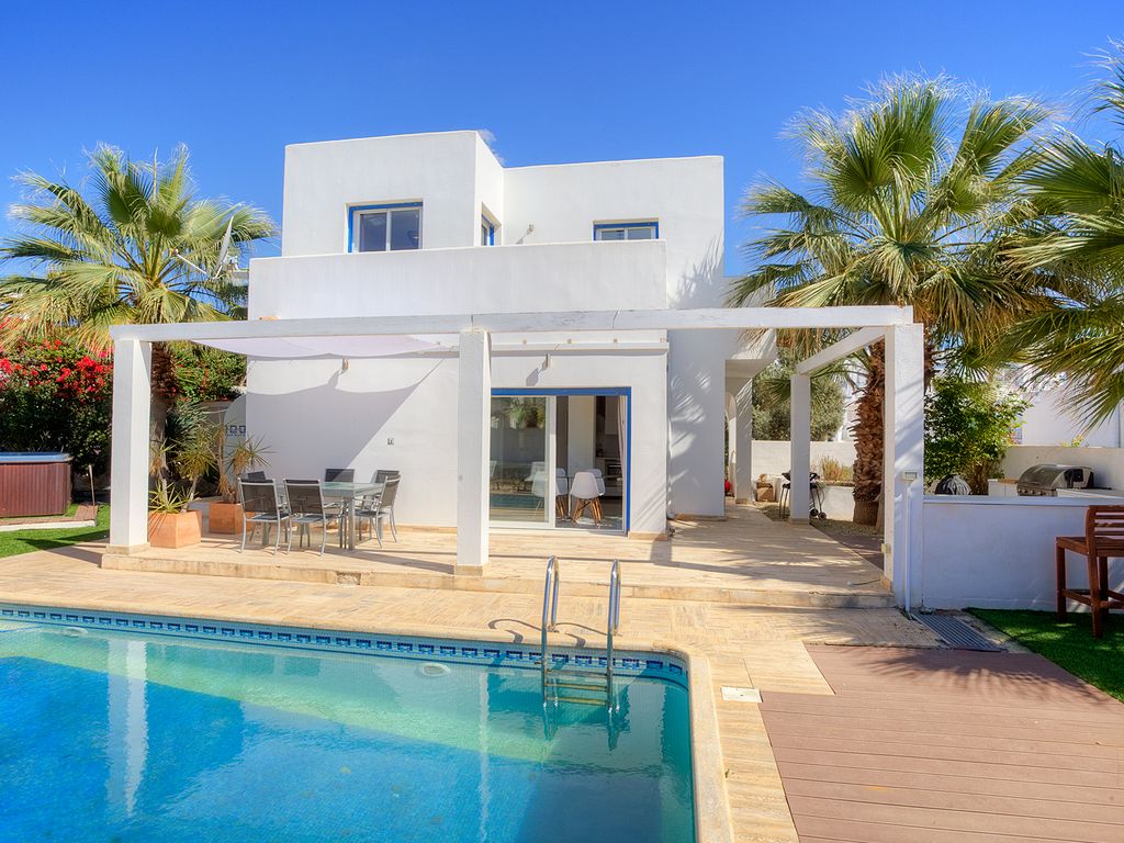 A modern villa exterior is shown, featuring a private swimming pool in the foreground. The patio is adorned with outdoor dining furniture and shaded by a pergola. Lush palm trees surround the property, and large glass doors invite view into the spacious interior.
