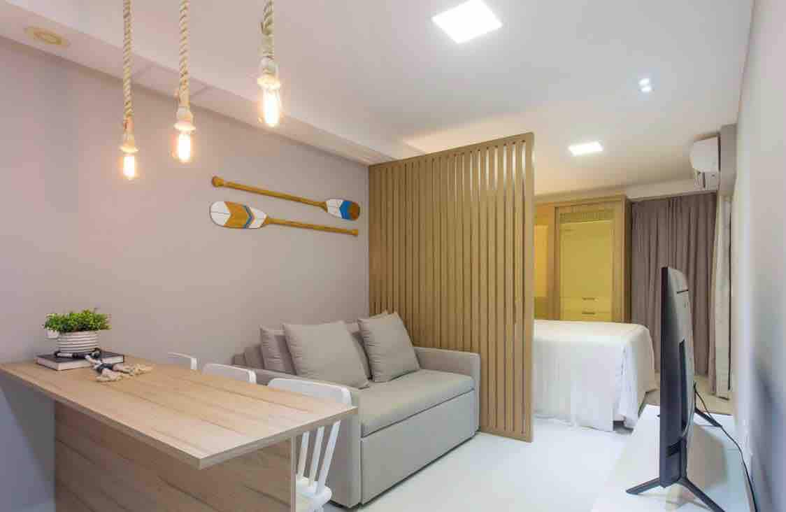 A modern living space features a comfortable sofa and a dining area with a light wood table. Decorative oars are mounted on the wall, and soft lighting is provided by hanging bulbs. A partition subtly separates the sleeping area, revealing a queen bed behind.