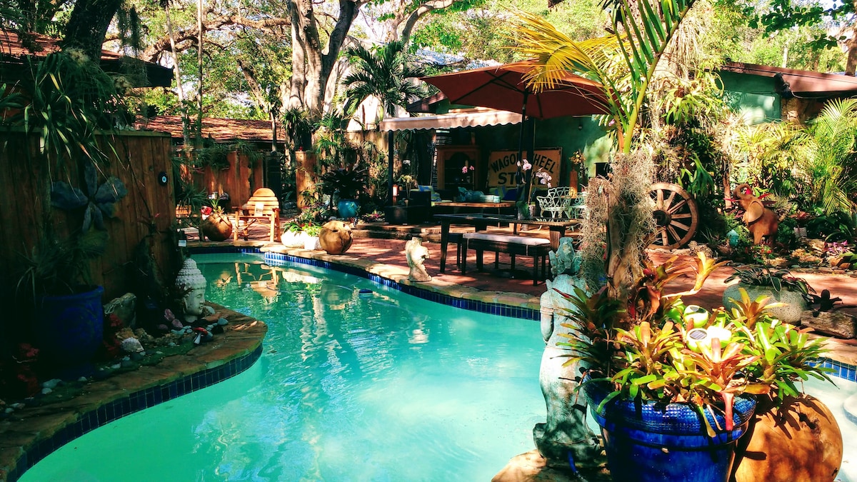 A serene swimming pool is framed by lush greenery and colorful plants. A shaded outdoor seating area is visible in the background, accompanied by a large umbrella. Natural stone pathways lead through the vibrant garden, enhancing the tranquil environment.