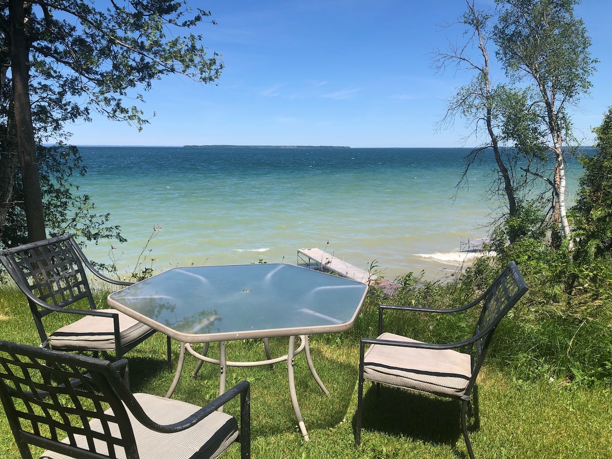 A table and four chairs are arranged on a grassy area overlooking the lake. The water's vibrant blue and green hues are visible, with distant land on the horizon. Trees frame the scene, contributing to the serene outdoor setting.