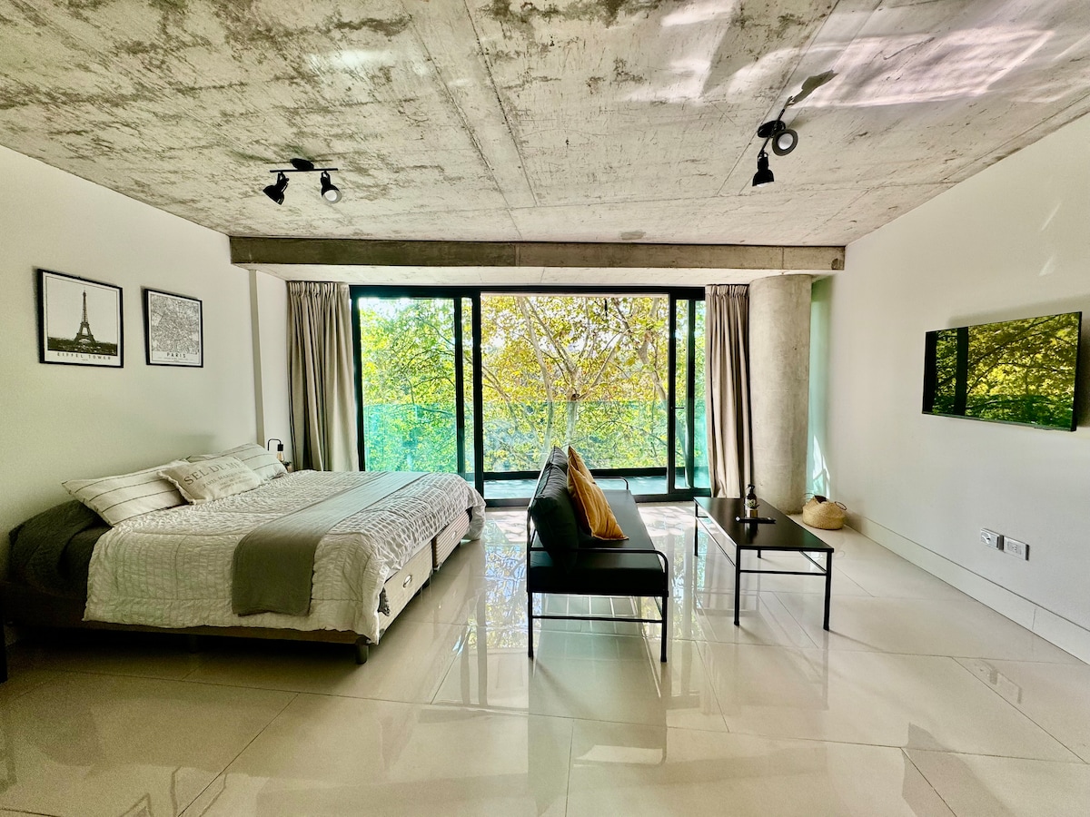 A spacious studio is shown with a large bed adorned in neutral tones. Floor-to-ceiling windows frame a vibrant green view of surrounding trees. A sleek TV hangs on the wall opposite, while a contemporary seating area is positioned beside a small black table.