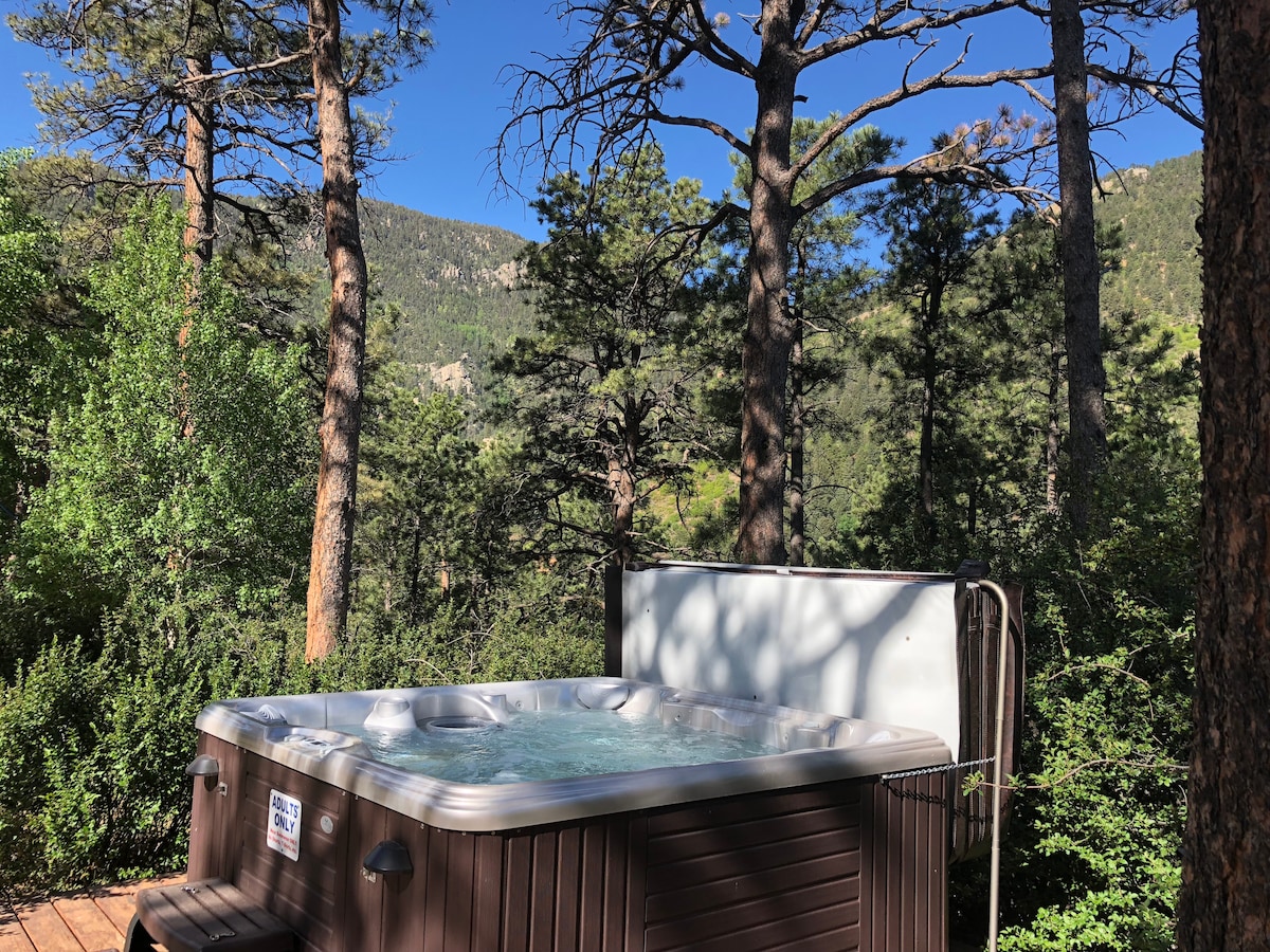 A private outdoor hot tub is surrounded by tall pine trees and lush greenery, offering views of the mountainous landscape. The serene setting invites relaxation, with clear blue skies overhead complementing the natural beauty of the area.